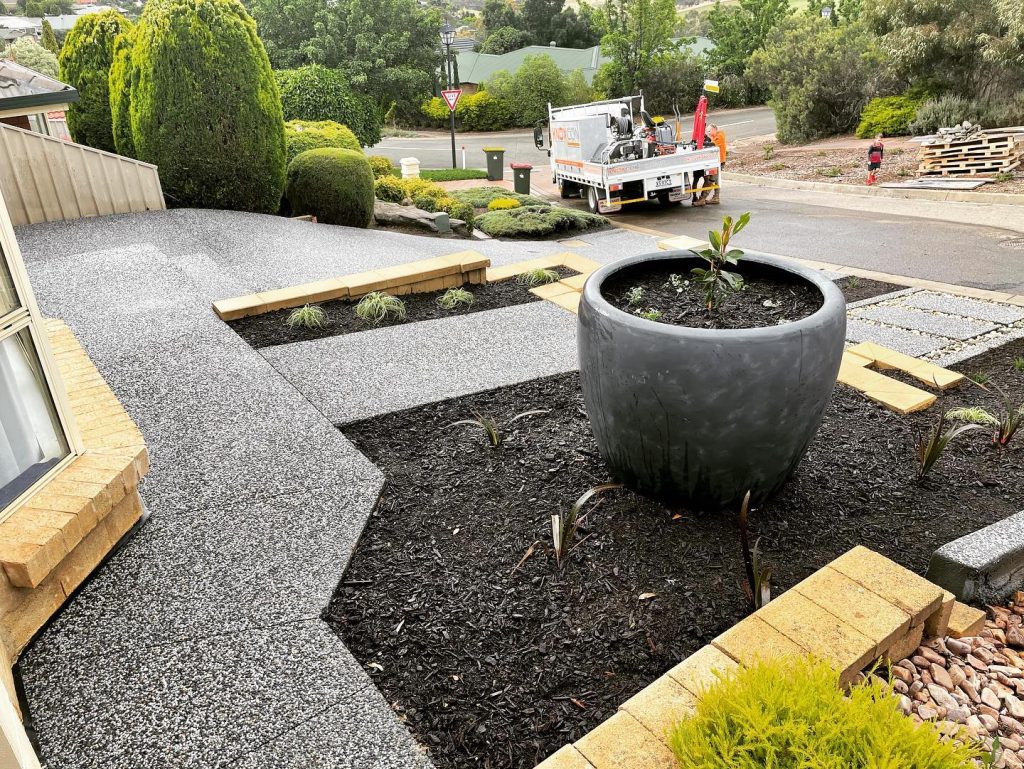 Front garden with exposed aggregate paths, black mulch beds and a large grey pot